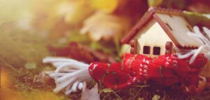 A wooden miniature house wrapped in a red, green and white scarf on the ground with fall leaves