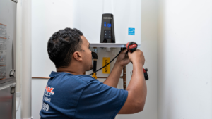A technician in a blue shirt holding a cordless drill in front of white tankless water heating unit