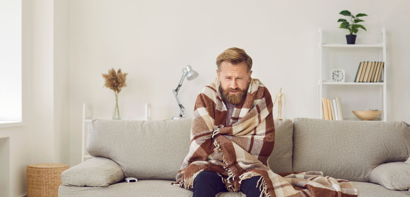 Sad bearded young guy wrapped in woolen plaid shivering while sitting on sofa in living room interior.